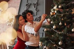 Garlands and silver balls. Romantic couple dressing up Christmas tree in the room with brown wall and fireplace photo