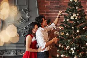 Romantic couple dressing up Christmas tree in the room with brown wall and fireplace photo