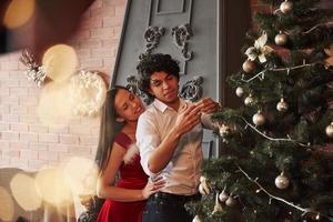 Nice interior. Romantic couple dressing up Christmas tree in the room with brown wall and fireplace photo