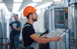 Using machine to program operations. Industrial worker indoors in factory. Young technician with orange hard hat photo