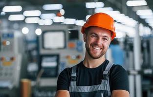 Smiling and happy employee. Portrait of industrial worker indoors in factory. Young technician with orange hard hat photo