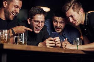 contenido divertido en el teléfono inteligente. tres amigos descansando en el pub con cerveza en las manos. teniendo una conversación foto