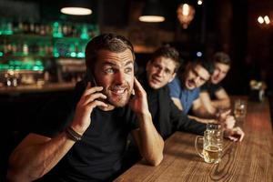 Man talks by the phone. Three sports fans in a bar watching soccer. With beer in hands