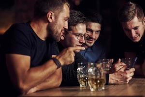 contenido divertido en el teléfono inteligente. tres amigos descansando en el pub con cerveza en las manos. teniendo una conversación foto