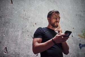 Messaging by using phone. Portrait of man in black shirt smoking on the background of old cracked wall photo