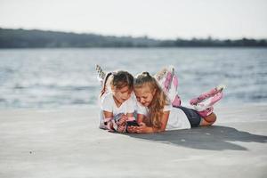 Using smartphone. Two little girls with roller skates outdoors near the lake at background photo