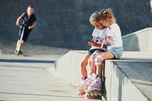 usando un teléfono inteligente. en la rampa para los deportes extremos. dos niñas pequeñas con patines al aire libre se divierten foto