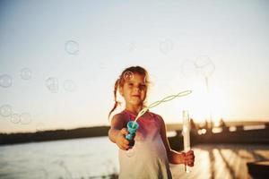 Evening time. Happy little girl playing with bubbles near the lake at park photo