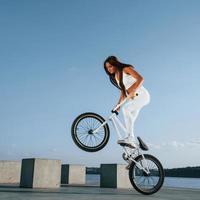 Doing stunts. Female rider is on the bike at daytime near the lake. Fitness woman in sportive clothes photo