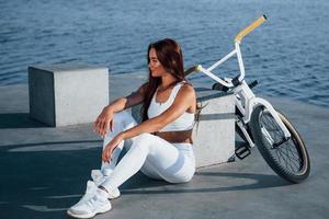 Near the bike. Fitness woman having a rest near the lake at daytime. Beautiful sunlight photo