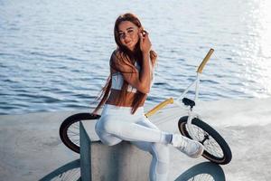 Near the bike. Fitness woman having a rest near the lake at daytime. Beautiful sunlight photo