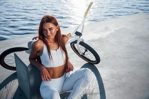 Near the bike. Fitness woman having a rest near the lake at daytime. Beautiful sunlight photo