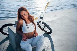 Near the bike. Fitness woman having a rest near the lake at daytime. Beautiful sunlight photo