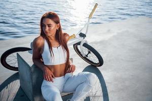 Near the bike. Fitness woman having a rest near the lake at daytime. Beautiful sunlight photo