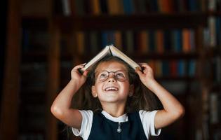 Cute little girl in glasses stands in the library full of books. Conception of education photo