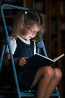 concentración en la lectura. niña de la escuela en la escalera en la biblioteca llena de libros. concepción de la educación foto