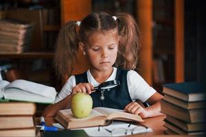 tiene lupa. linda niña con coletas está en la biblioteca. manzana en los libros foto