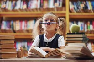 Pencil on lips. Cute little girl with pigtails is in the library. Apple on the books photo