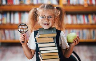 tiene lupa. linda niña con coletas está en la biblioteca. manzana en los libros foto