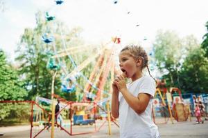 una niña linda come helado en el parque durante el día cerca de las atracciones foto