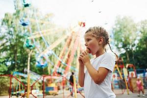 una niña linda come helado en el parque durante el día cerca de las atracciones foto