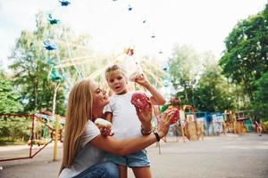 comiendo donas una niña alegre en patines y su madre se divierten juntos en el parque cerca de las atracciones foto