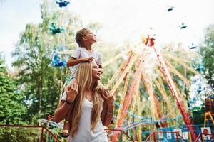 Daughter sits on the shoulders. Cheerful little girl her mother have a good time in the park together near attractions photo