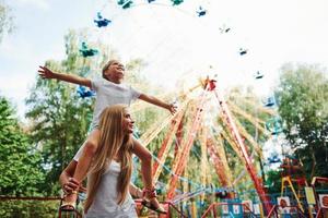 hija se sienta en los hombros. niña alegre su madre se divierten juntos en el parque cerca de las atracciones foto