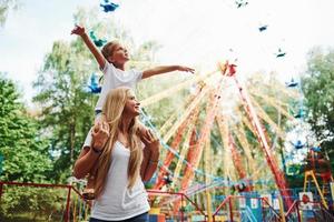 Daughter sits on the shoulders. Cheerful little girl her mother have a good time in the park together near attractions photo