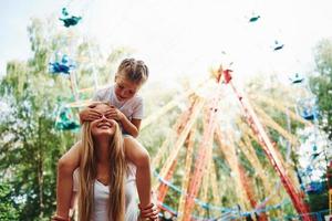 hija se sienta en los hombros. niña alegre su madre se divierten juntos en el parque cerca de las atracciones foto