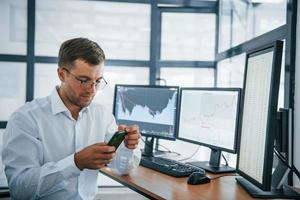 Using smartphone. Young businessman in formal clothes is in office with multiple screens. Conception of exchange and money photo