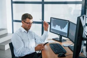 Using smartphone. Young businessman in formal clothes is in office with multiple screens. Conception of exchange and money photo