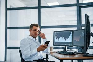 Using smartphone. Young businessman in formal clothes is in office with multiple screens. Conception of exchange and money photo