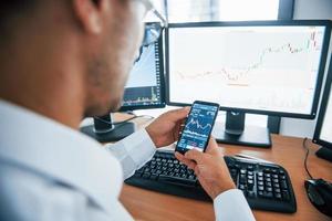 Using smartphone. Young businessman in formal clothes is in office with multiple screens. Conception of exchange and money photo