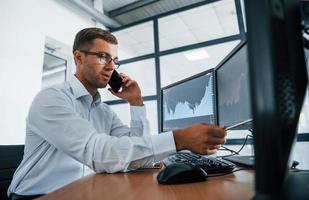 Having conversation by the phone. Looks at the graphs. Young businessman in formal clothes is in office with multiple screens photo