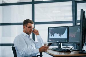 Using smartphone. Young businessman in formal clothes is in office with multiple screens. Conception of exchange and money photo