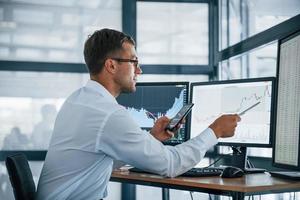 With phone and pen. Analysing graphs. Young businessman in formal clothes is in office with multiple screens. Conception of exchange and money photo