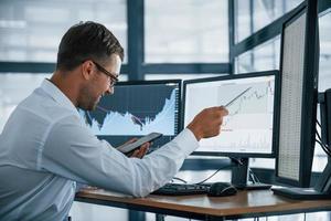 With phone and pen. Analysing graphs. Young businessman in formal clothes is in office with multiple screens. Conception of exchange and money photo