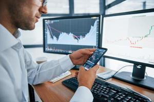 Using smartphone. Young businessman in formal clothes is in office with multiple screens. Conception of exchange and money photo