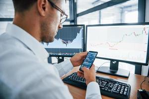 Using smartphone. Young businessman in formal clothes is in office with multiple screens. Conception of exchange and money photo