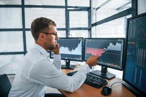Having conversation by the phone. Looks at the graphs. Young businessman in formal clothes is in office with multiple screens photo