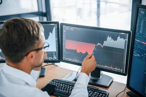 Looks at the graphs. Young businessman in formal clothes is in office with multiple screens. Conception of exchange and money photo