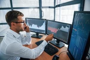 Having conversation by the phone. Looks at the graphs. Young businessman in formal clothes is in office with multiple screens photo