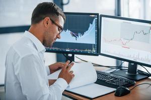 Works with documents. Young businessman in formal clothes is in office with multiple screens. Conception of exchange and money photo