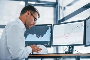 Works with documents. Young businessman in formal clothes is in office with multiple screens. Conception of exchange and money photo
