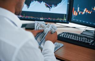 Calculating money. Young businessman in formal clothes is in office with multiple screens. Conception of exchange and money photo