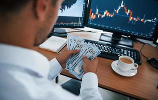 Calculating money. Young businessman in formal clothes is in office with multiple screens. Conception of exchange and money photo