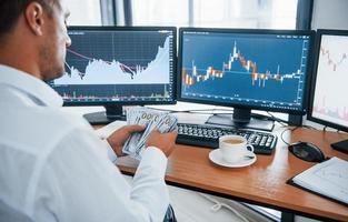 Calculating money. Young businessman in formal clothes is in office with multiple screens. Conception of exchange and money photo