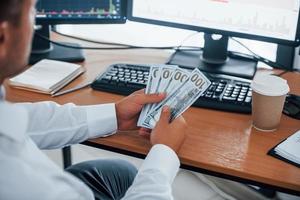 Calculating money. Young businessman in formal clothes is in office with multiple screens. Conception of exchange and money photo