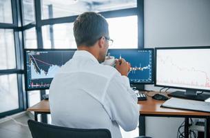 Holds cup of drink. Analysing information. Young businessman in formal clothes is in office with multiple screens. Conception of exchange and money photo